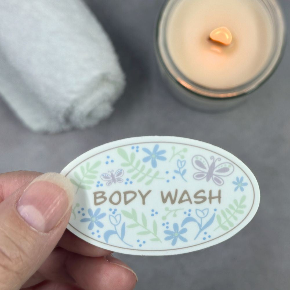 Hand holding a 'BODY WASH' label with floral designs on a gray surface with a candle and towel.